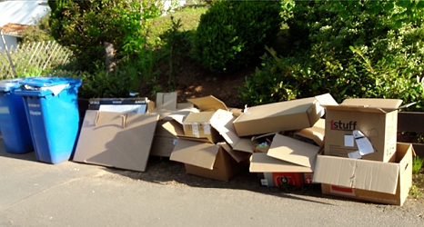 Das Foto zeigt eine ungeordnete Reihe aufeinander gelegter Kartons neben Altpapiertonnen. © DBM